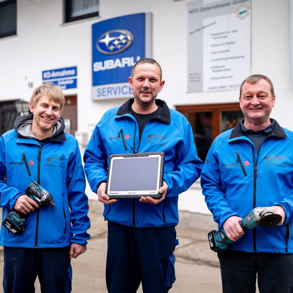 Die drei Meister vor der KFZ-Werkstatt Zehendmaier. Drei KFZ-Meister in blauen Jacken halten Werkzeuge und ein Tablet vor der KFZ-Werkstatt.
