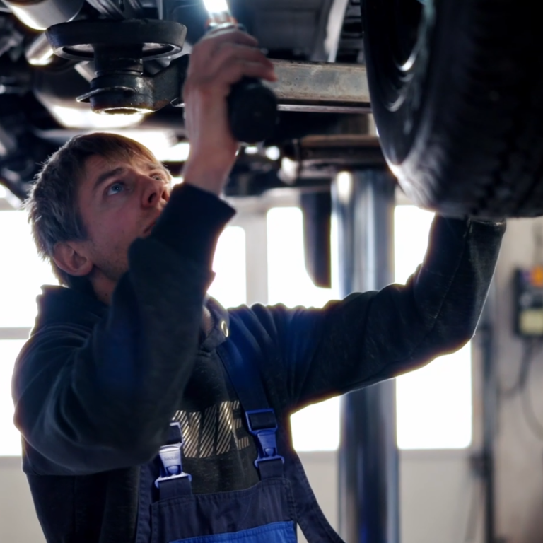 Mechanikermeister inspiziert den Unterboden. Junger Mechanikermeister arbeitet unter einem Auto mit einem Werkzeug.